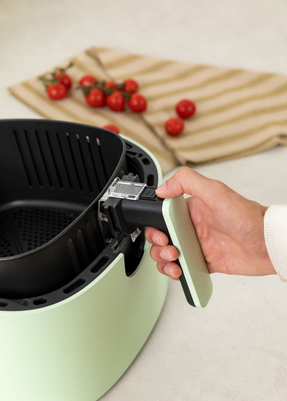 Ręka trzyma uchwyt miętowej frytkownicy. Czerwone pomidory cherry i pasiasta ściereczka w tle.