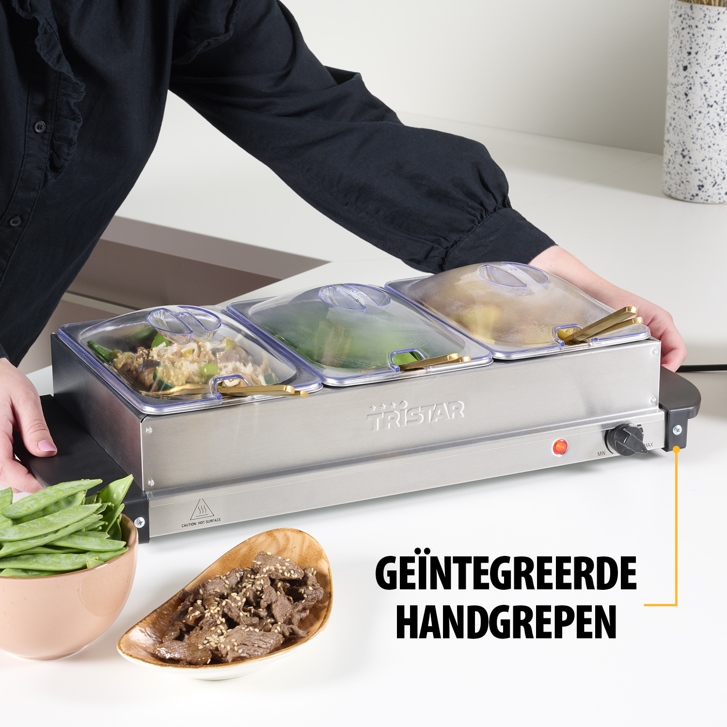Eine Person hält einen Speisenwärmer mit drei Fächern. Schüsseln mit Essen stehen auf der Theke. Der Wärmer trägt den Markennamen Tristar.