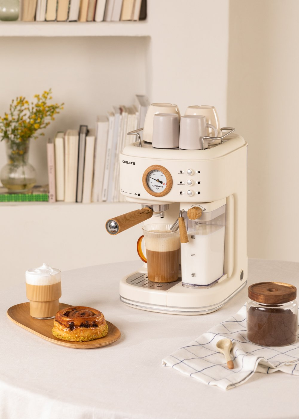 Una máquina de espresso crema con café, pasteles y tazas en una mesa blanca. Estantería y flores al fondo.