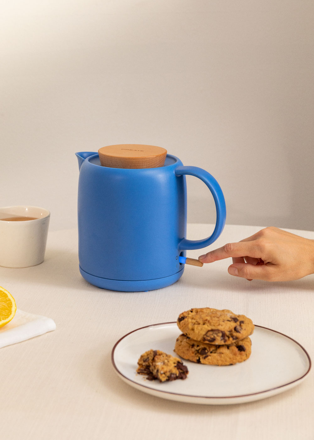 Una tetera azul con tapa de madera, una mano encendiéndola, galletas en un plato y una taza de té en una mesa.