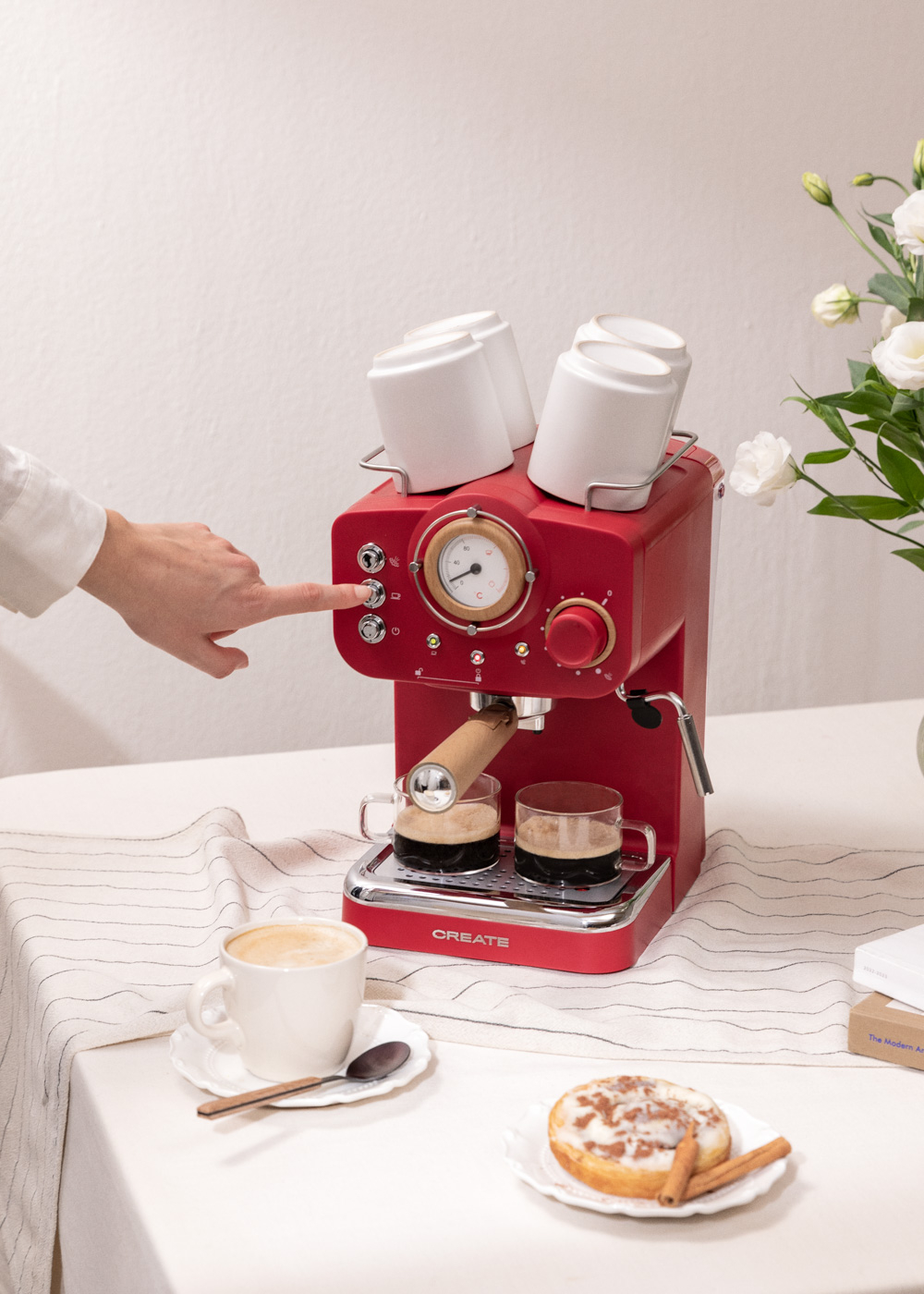 Una máquina de espresso roja, dos tazas de café, un latte y un pastel en una mesa. Una mano presiona un botón.