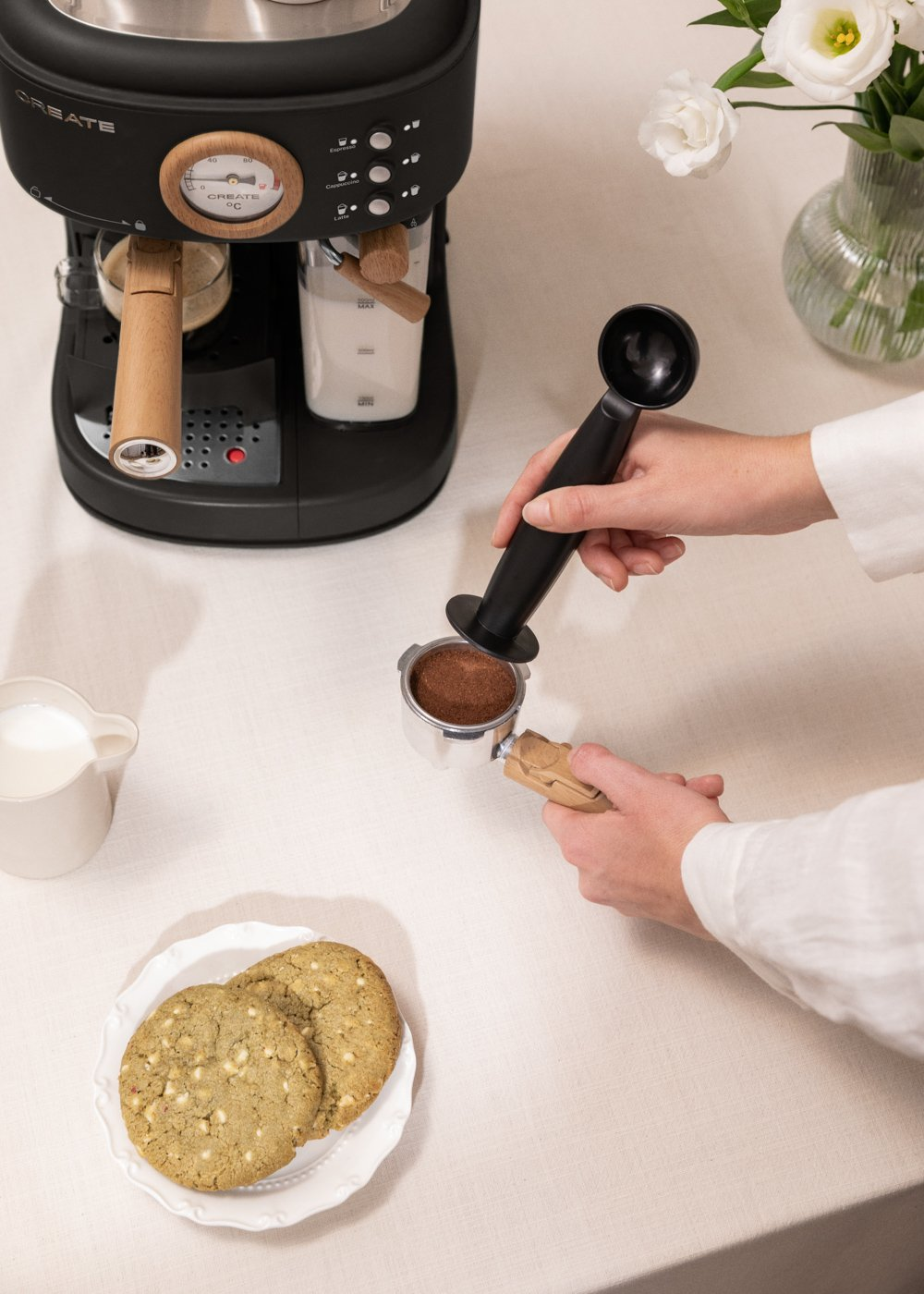 Una persona prepara café con una máquina de espresso. Hay galletas y leche en la mesa.