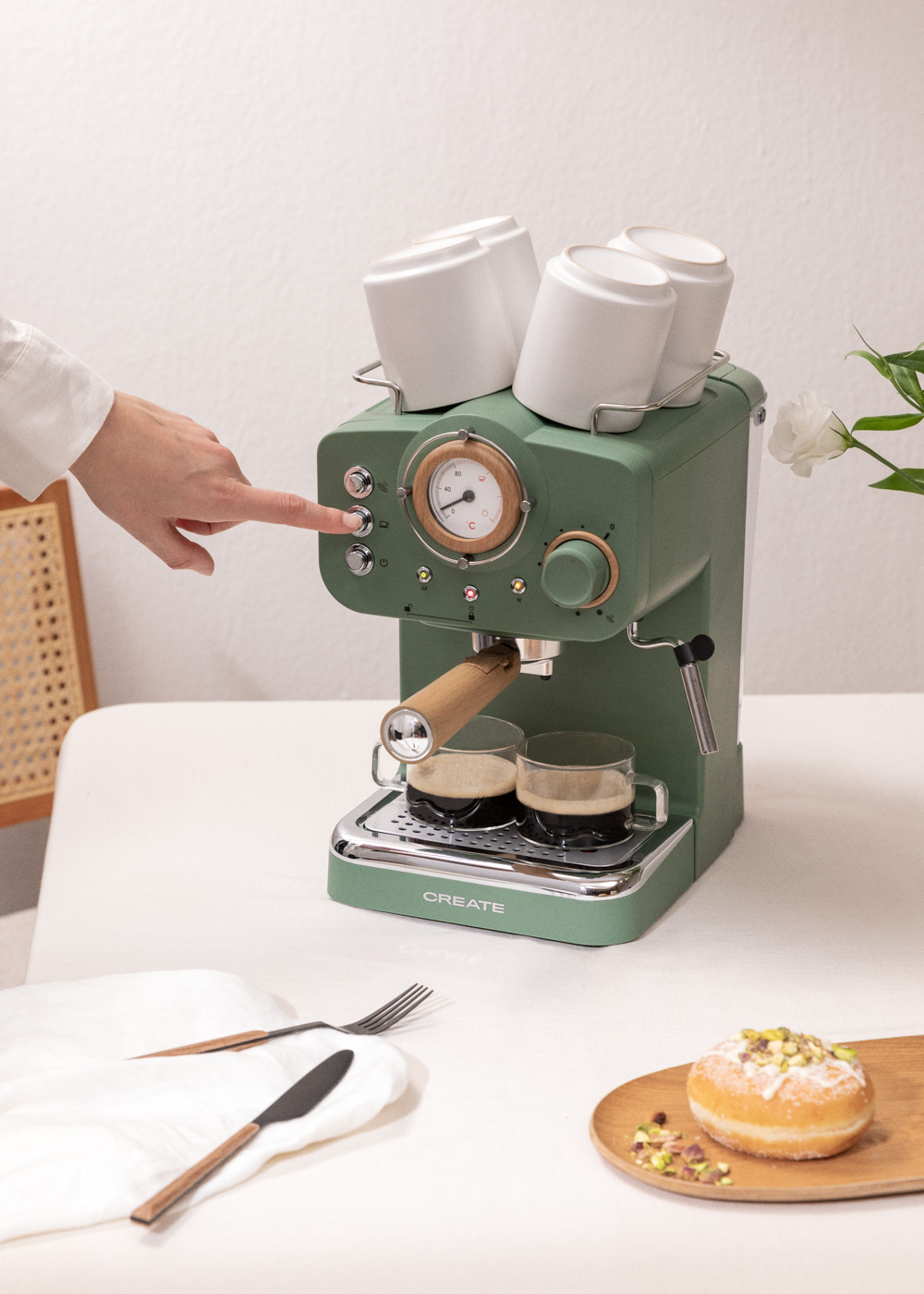 Una mano presiona un botón en una máquina de espresso verde. Dos tazas con café abajo. Tazas blancas arriba. Donut y utensilios cerca.