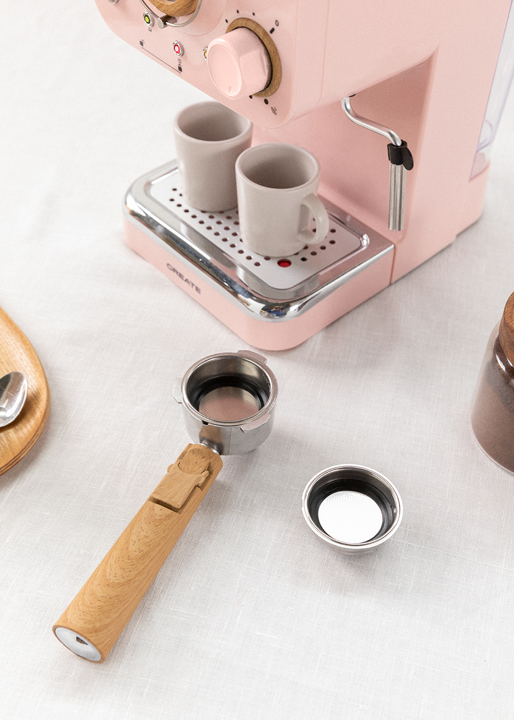 Una máquina de espresso rosa con dos tazas pequeñas, un portafiltro y un filtro sobre una superficie blanca.