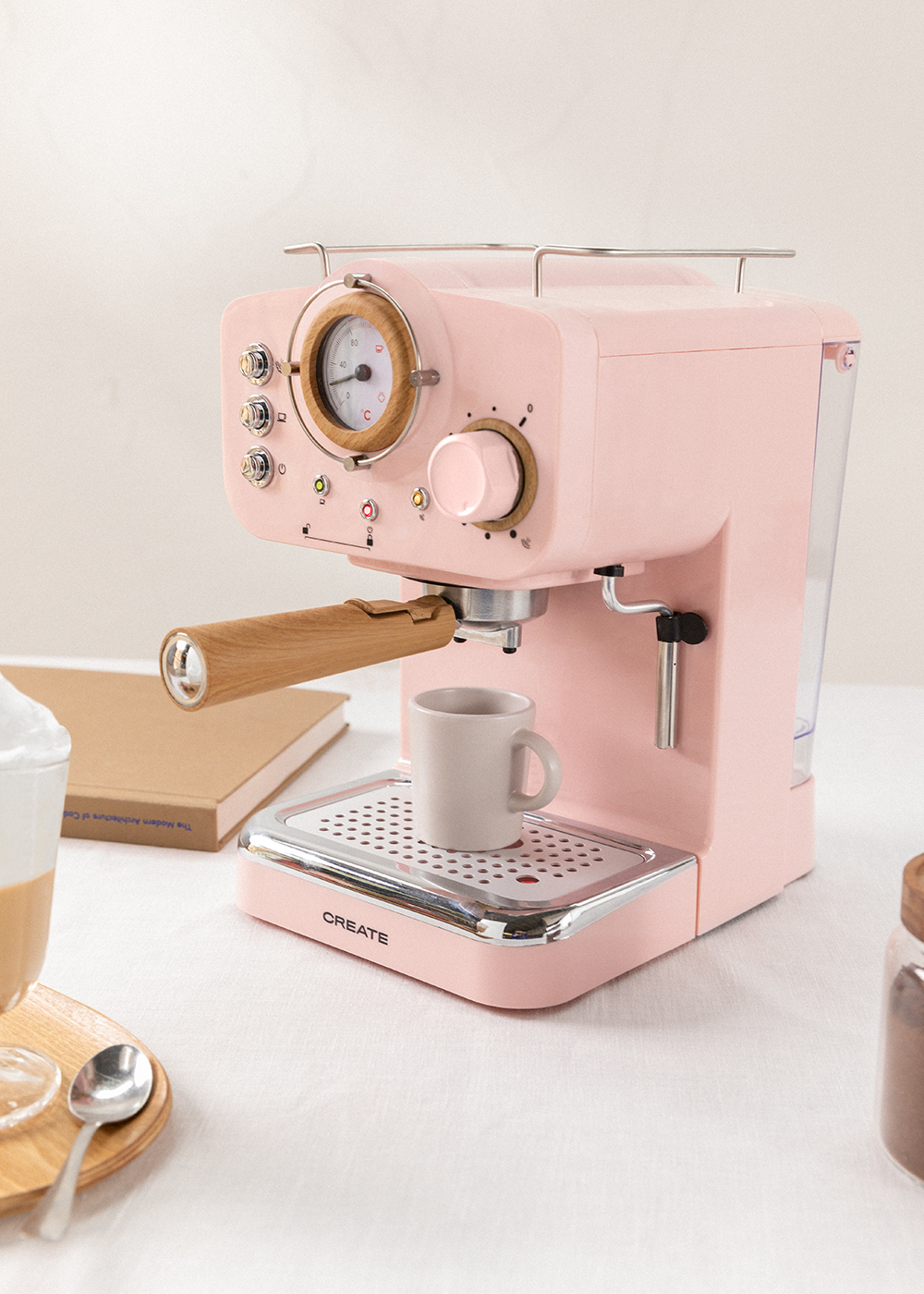 Una máquina de espresso rosa con mango de madera y taza. También se ven un libro, una bandeja con una bebida y un frasco.