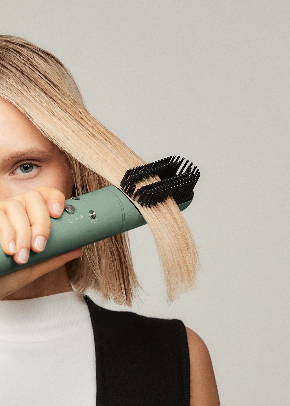 Una mujer usa una herramienta verde para peinar el cabello rubio. La herramienta se sostiene cerca de su rostro.