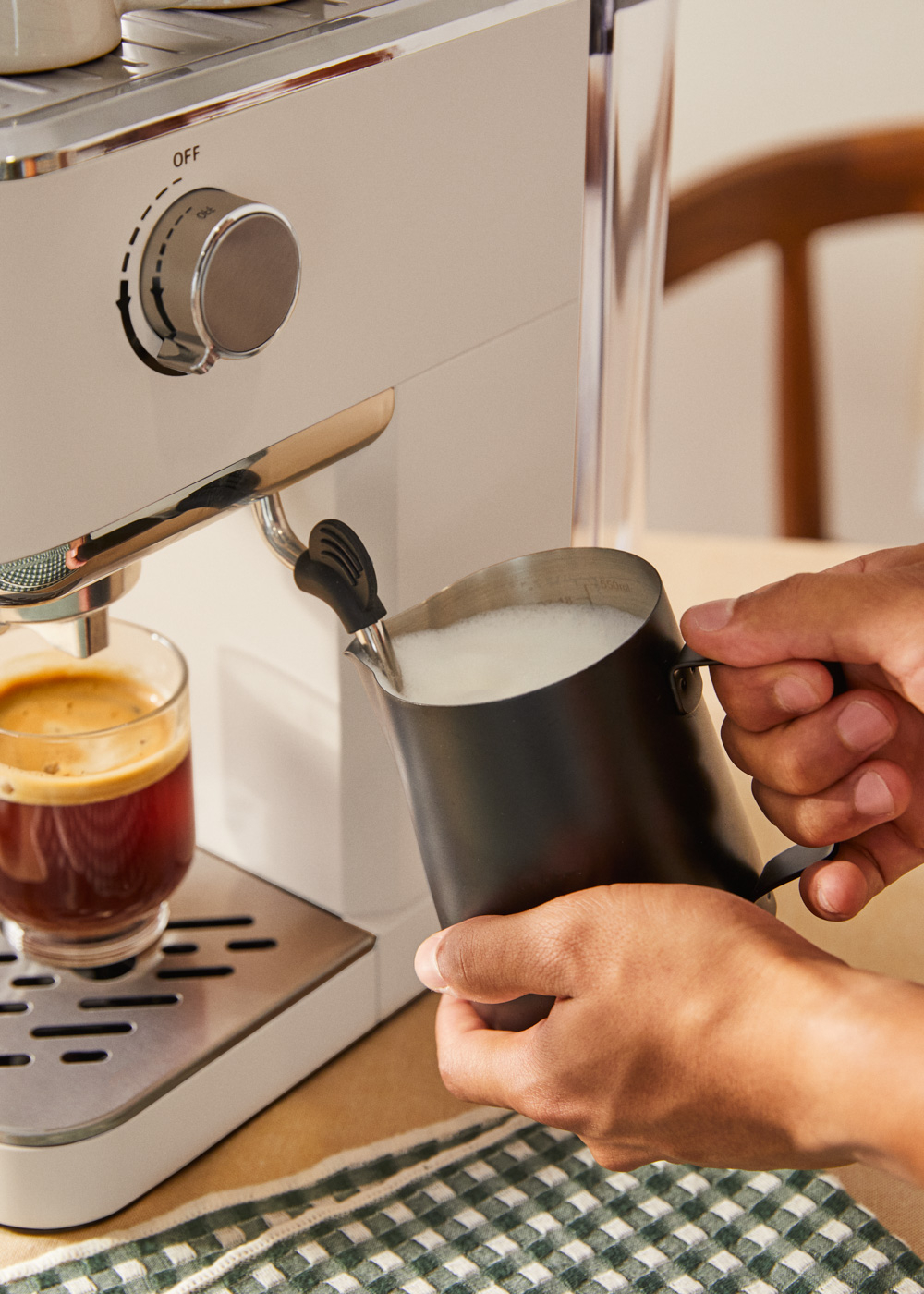 Una persona vierte leche espumada de una jarra negra en un vaso de espresso. Una máquina de espresso blanca está en el fondo.