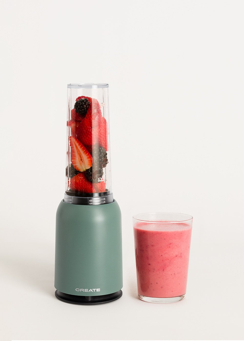 Una licuadora con fresas y moras, junto a un vaso de batido rosa sobre un fondo blanco.