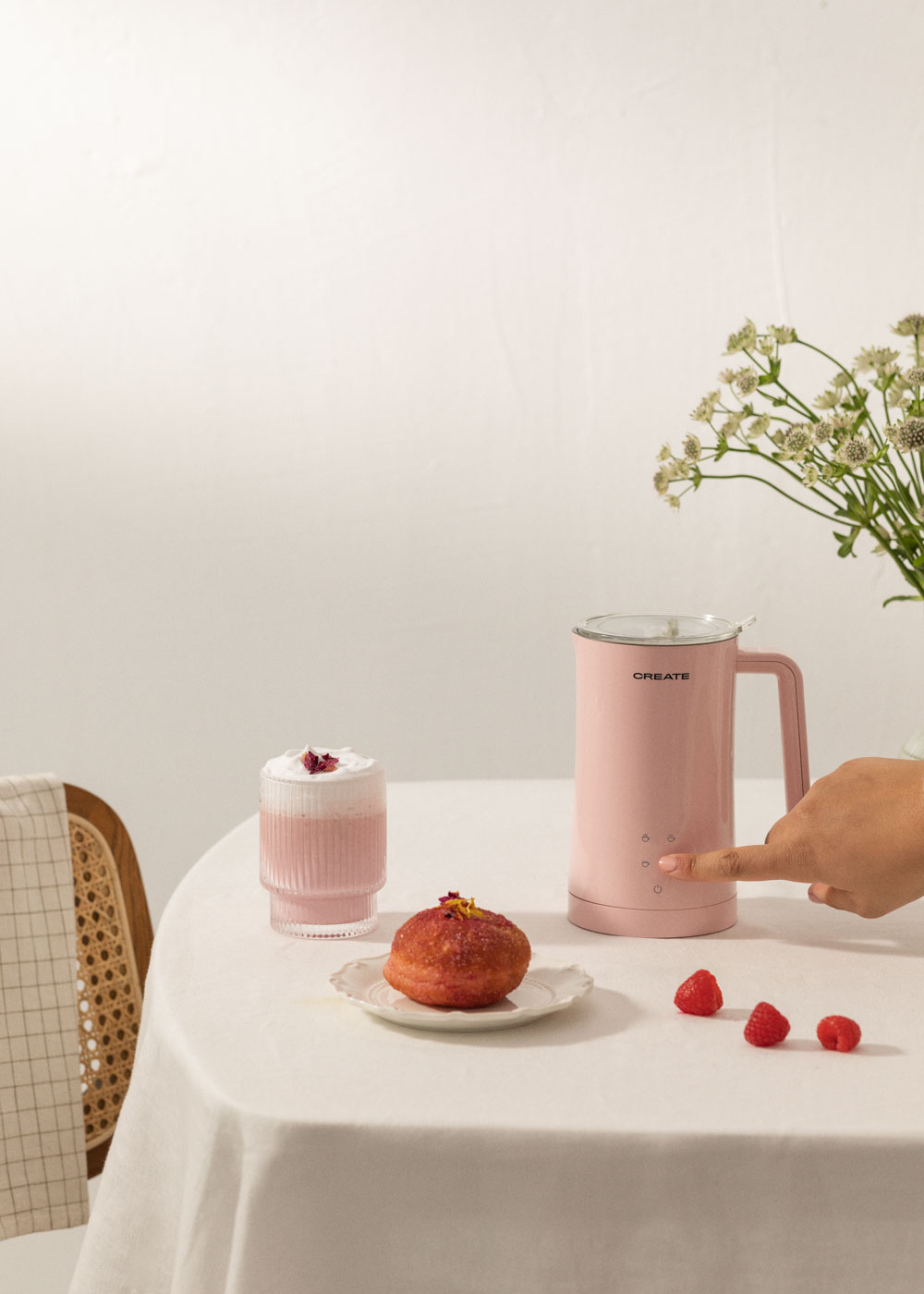 Una espumadora de leche rosa, una bebida, una rosquilla en un plato y tres frambuesas sobre un mantel blanco.