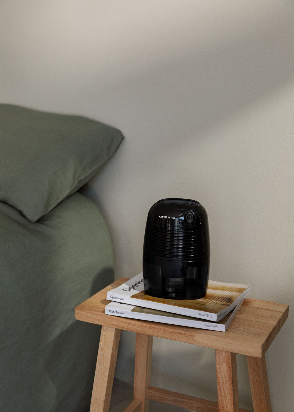 Un purificador de aire negro está sobre revistas en un taburete de madera. A la izquierda hay una almohada verde. El fondo es una pared beige.