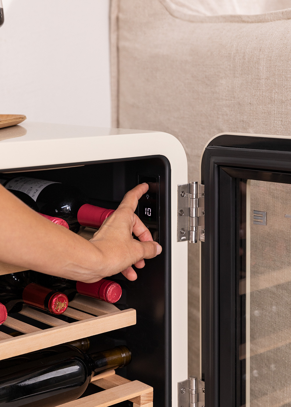 Una mano ajusta la temperatura de una vinoteca. Se ven botellas de vino tinto. Sofá beige al fondo.