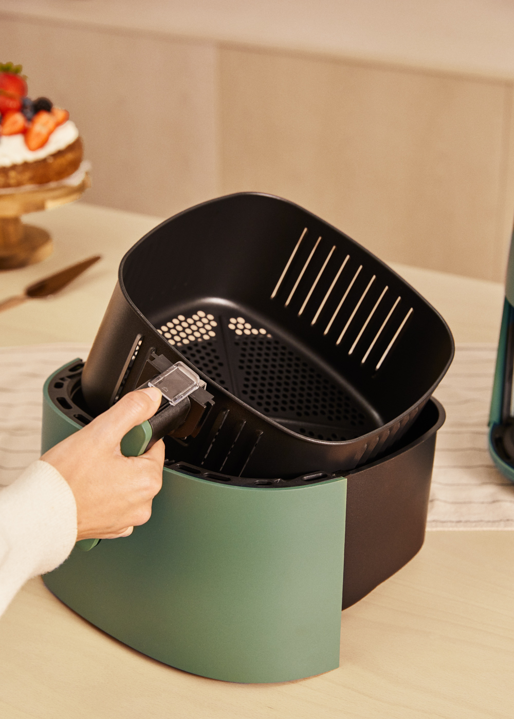 Osoba trzyma czarny kosz zielonej frytkownicy. W tle stoi ciasto z jagodami.