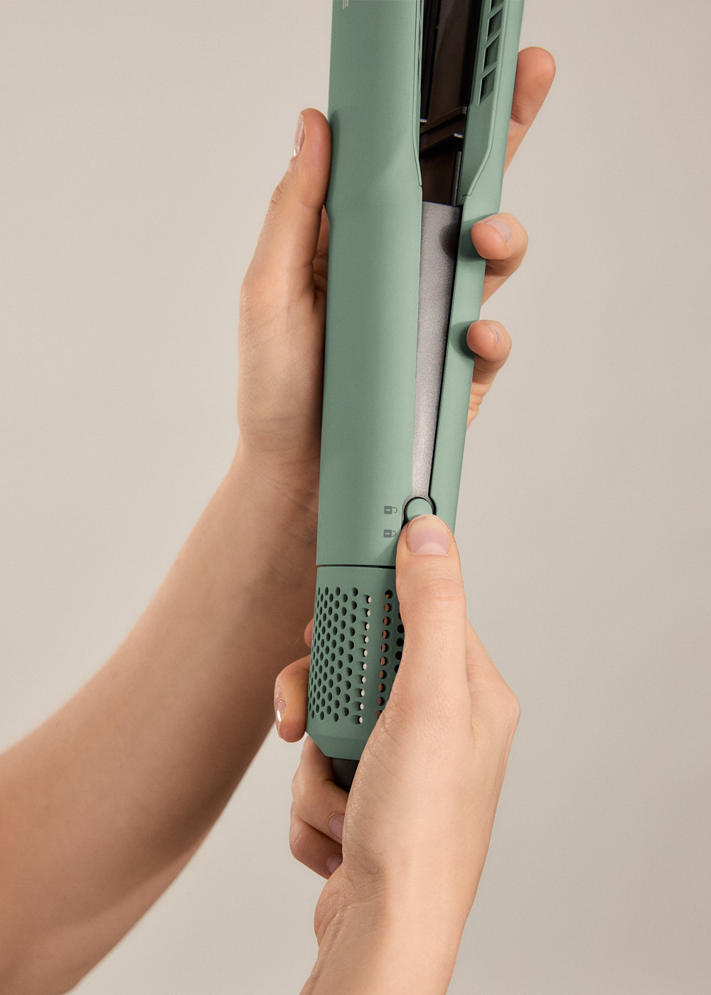 Manos sosteniendo una plancha de pelo verde. Una mano presiona un botón. El fondo es de color beige claro.