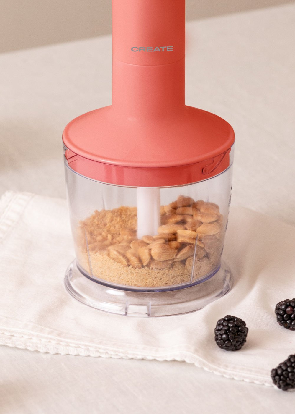 Un procesador de alimentos CREATE rosa con nueces trituradas y moras sobre tela blanca.