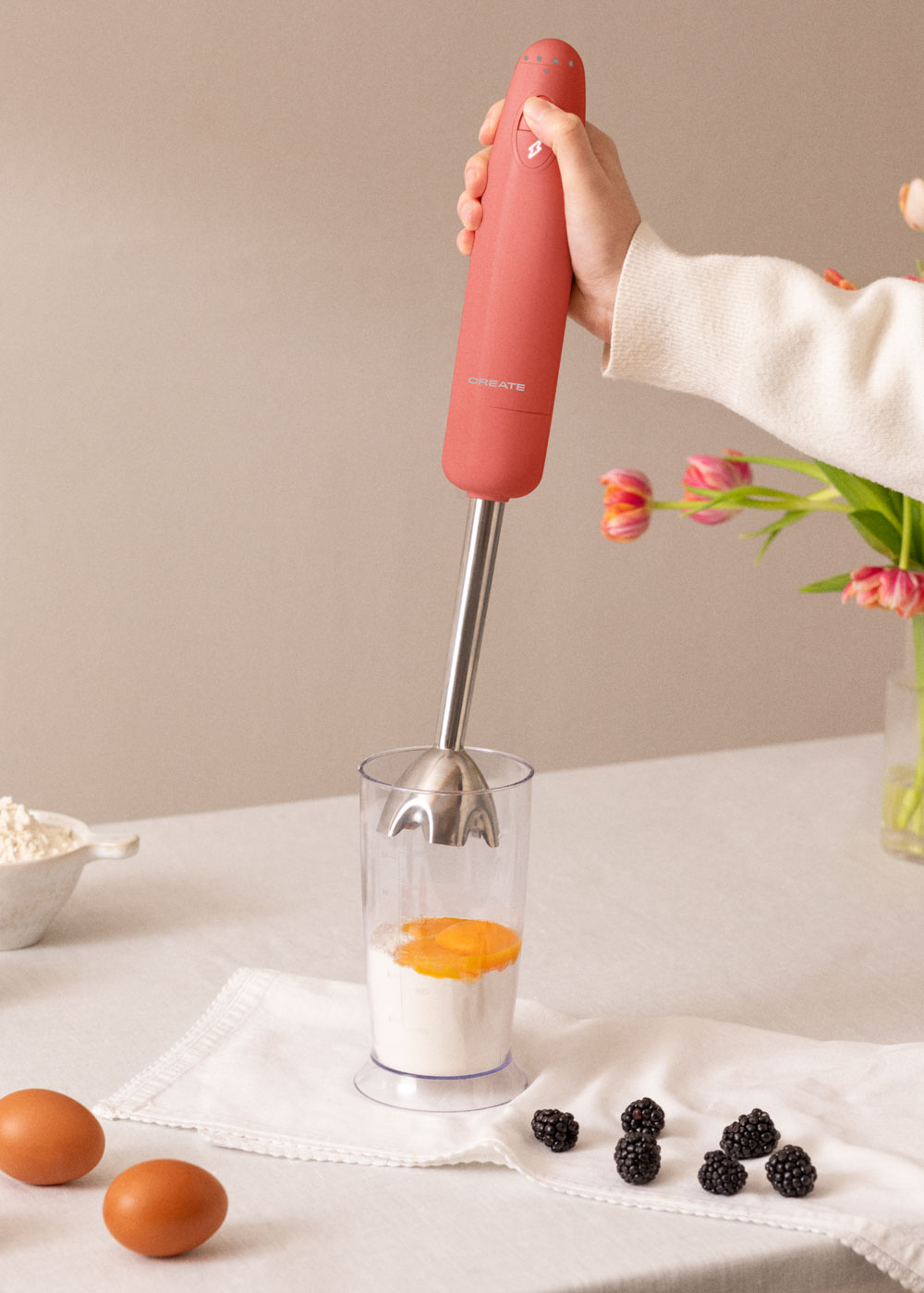 Una mano sostiene una batidora rosa sobre un vaso con yema de huevo y harina. Dos huevos y moras en la mesa blanca. Flores en el fondo.