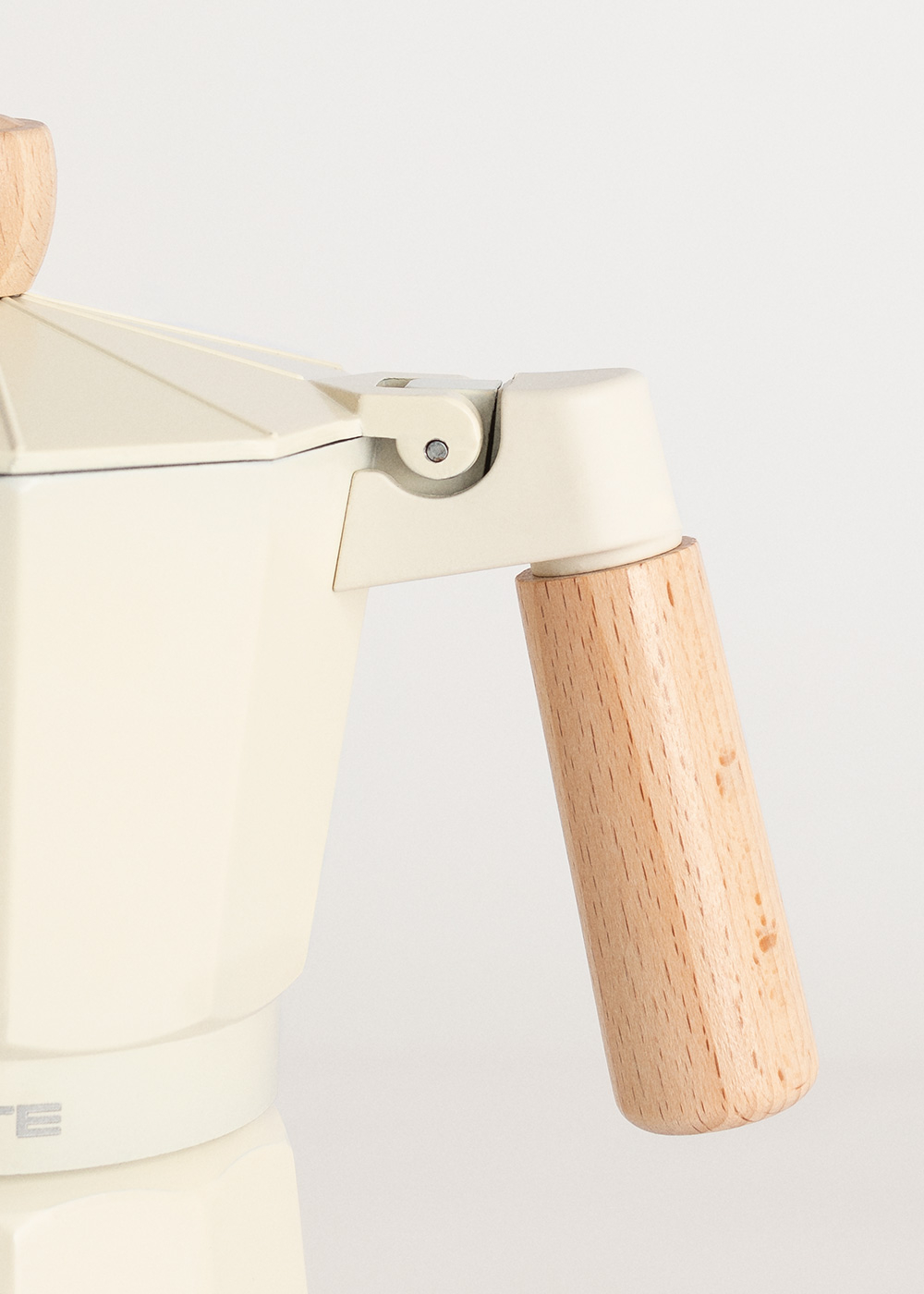 Primer plano de una cafetera crema con un asa de madera. El asa es cilíndrica y el fondo es blanco.