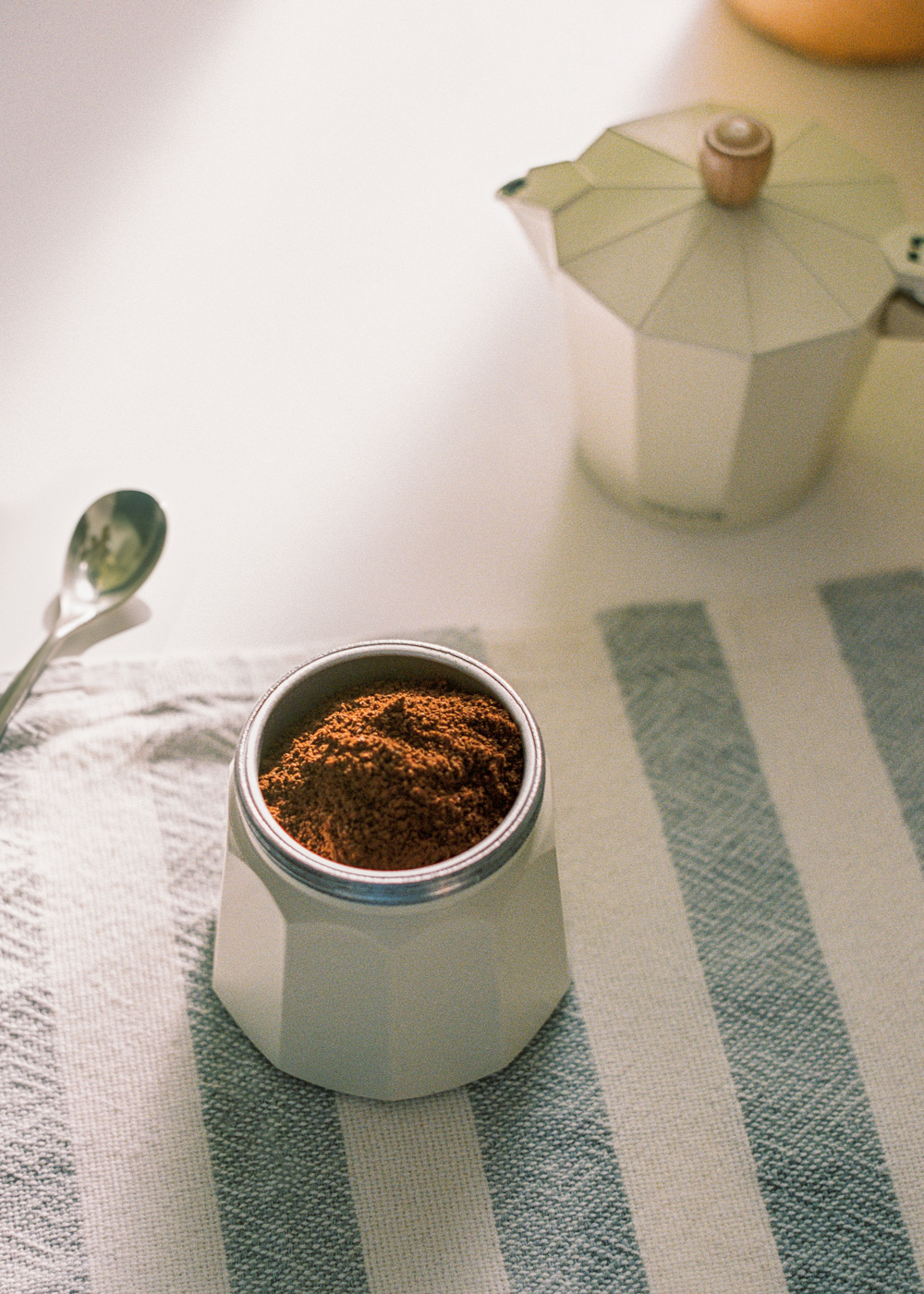 Una cafetera con café molido, una cuchara y una cafetera italiana sobre un paño a rayas.