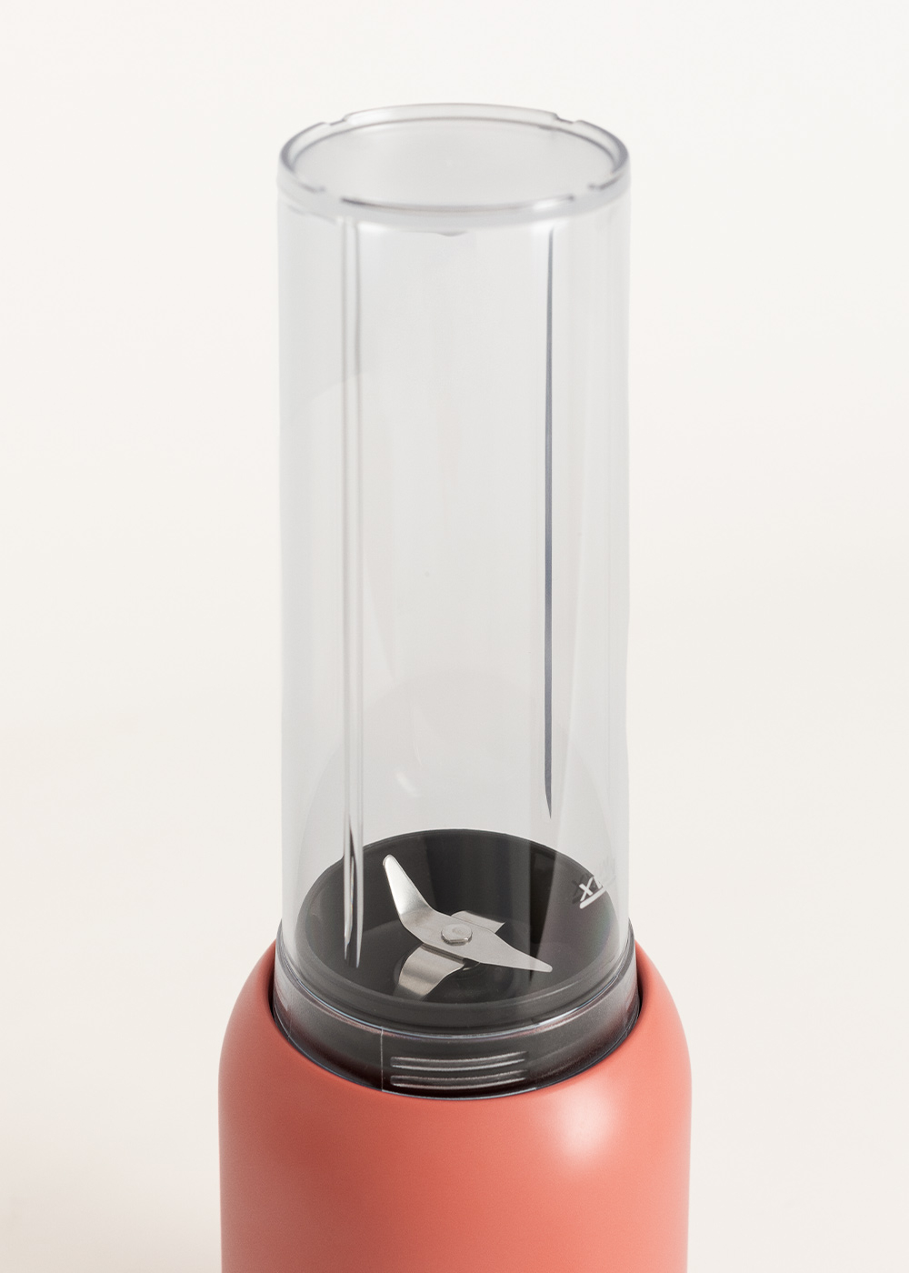 Primer plano de una licuadora. Tiene un recipiente de plástico transparente con una cuchilla en la parte inferior. La base es coral.