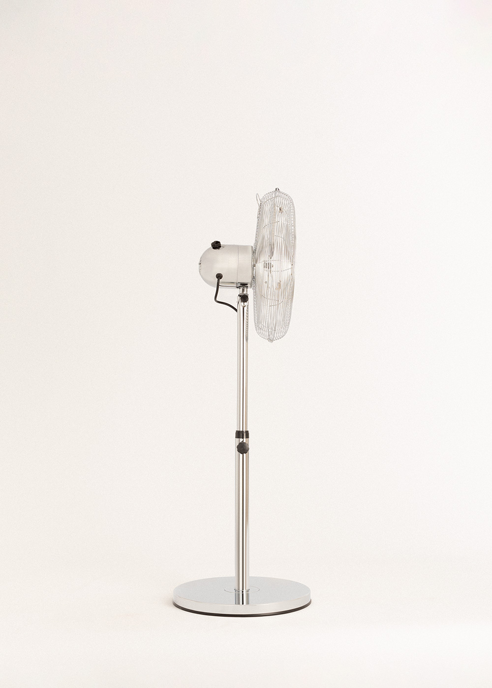 Un ventilador de pie plateado sobre un fondo blanco. El ventilador está encendido, inclinado hacia la derecha. La base es circular.