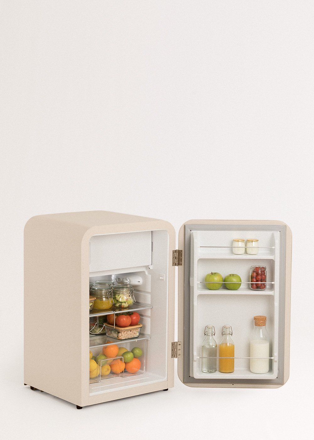 Un mini-refrigerador beige con la puerta abierta, lleno de frutas, verduras, frascos, botellas y productos lácteos. Fondo blanco.
