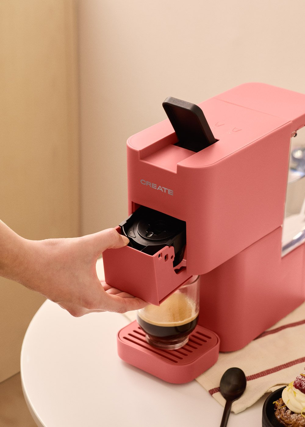 Una cafetera rosa con un cajón que se extrae. Debajo hay un vaso con café. Una mano tira del cajón.