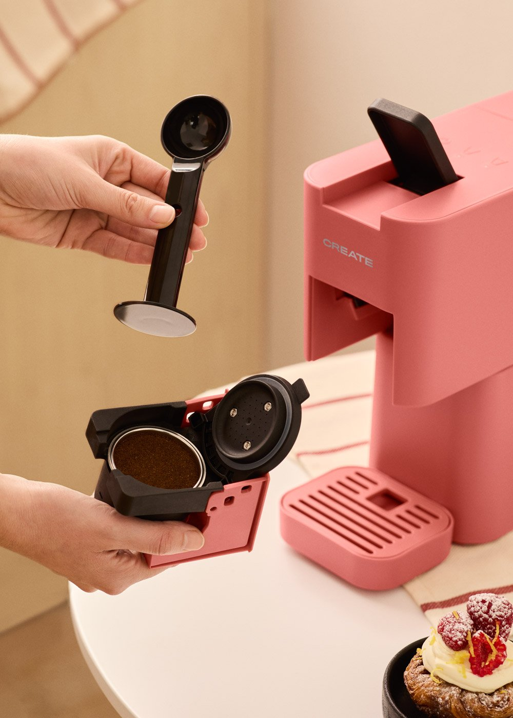 Una persona usa una cafetera. Es rosa, con una cuchara y una cápsula de café. Un pastel con crema y frambuesas está en la mesa.