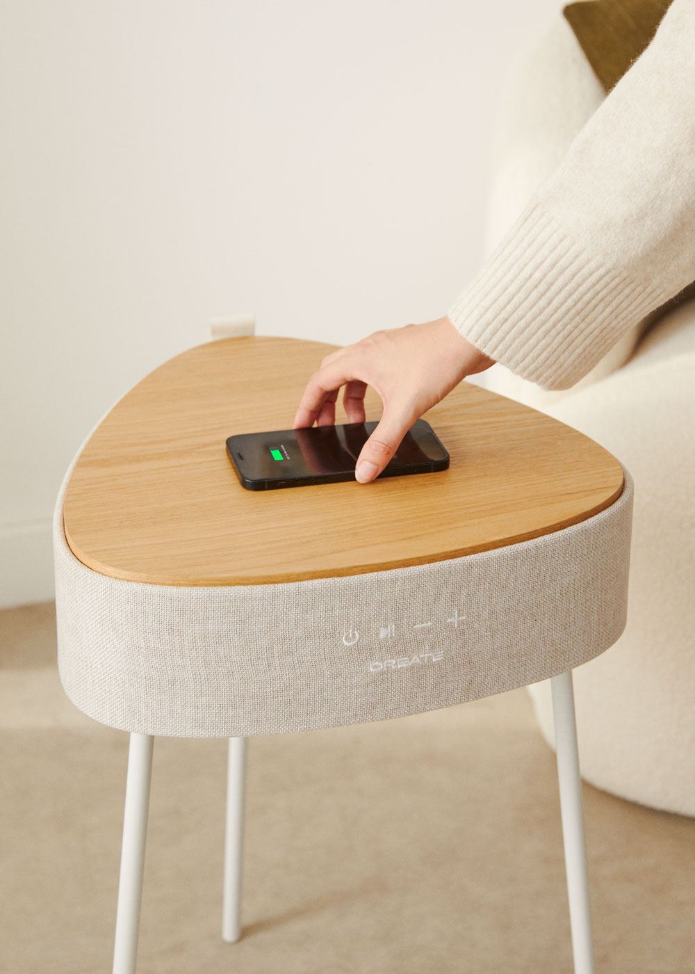 Una mano coloca un teléfono cargando sobre una mesa de madera. La mesa tiene una base de altavoz y patas blancas. Se ve un suéter color crema.