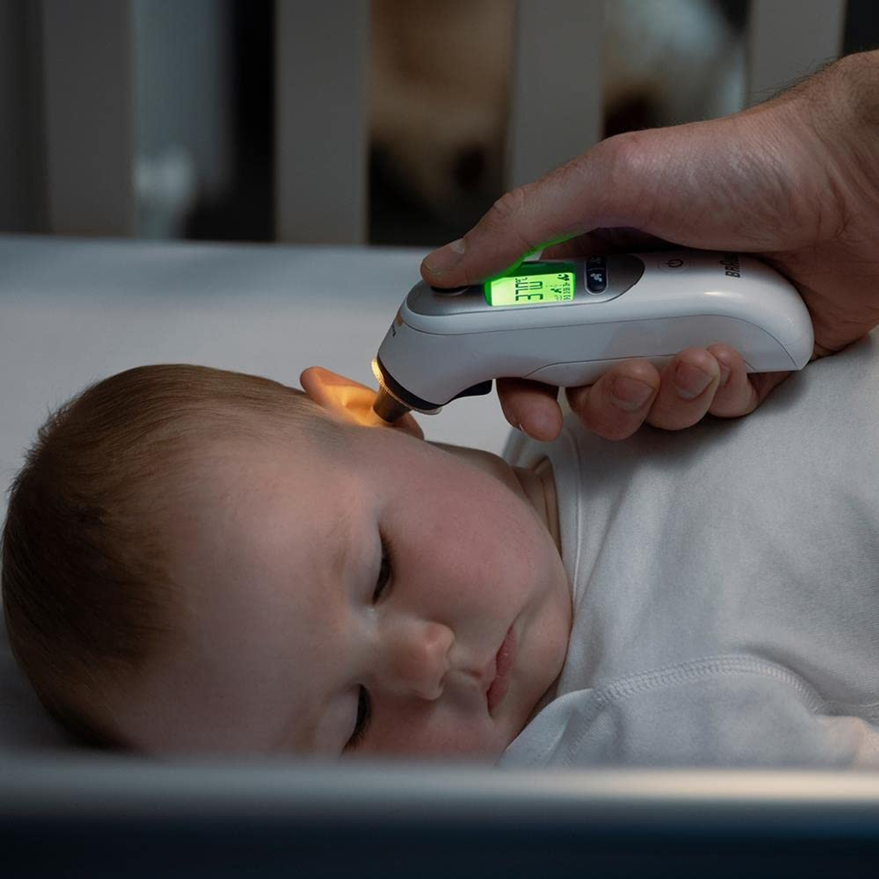 Eine Hand hält ein Thermometer an das Ohr eines Babys. Das Thermometer zeigt eine Temperatur an. Das Baby liegt in einem Kinderbett.