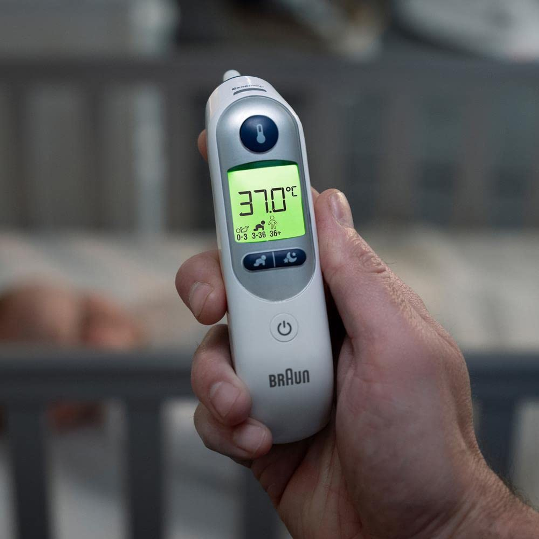 Eine Hand hält ein Braun-Thermometer, das 37,0 °C anzeigt. Im verschwommenen Hintergrund ist ein Babybett.