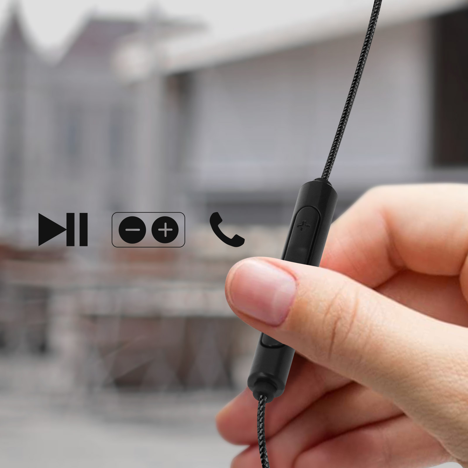 Una mano tiene un controllo nero per cuffie. Sono visibili icone di riproduzione, volume e telefono. Un edificio sfocato sullo sfondo.