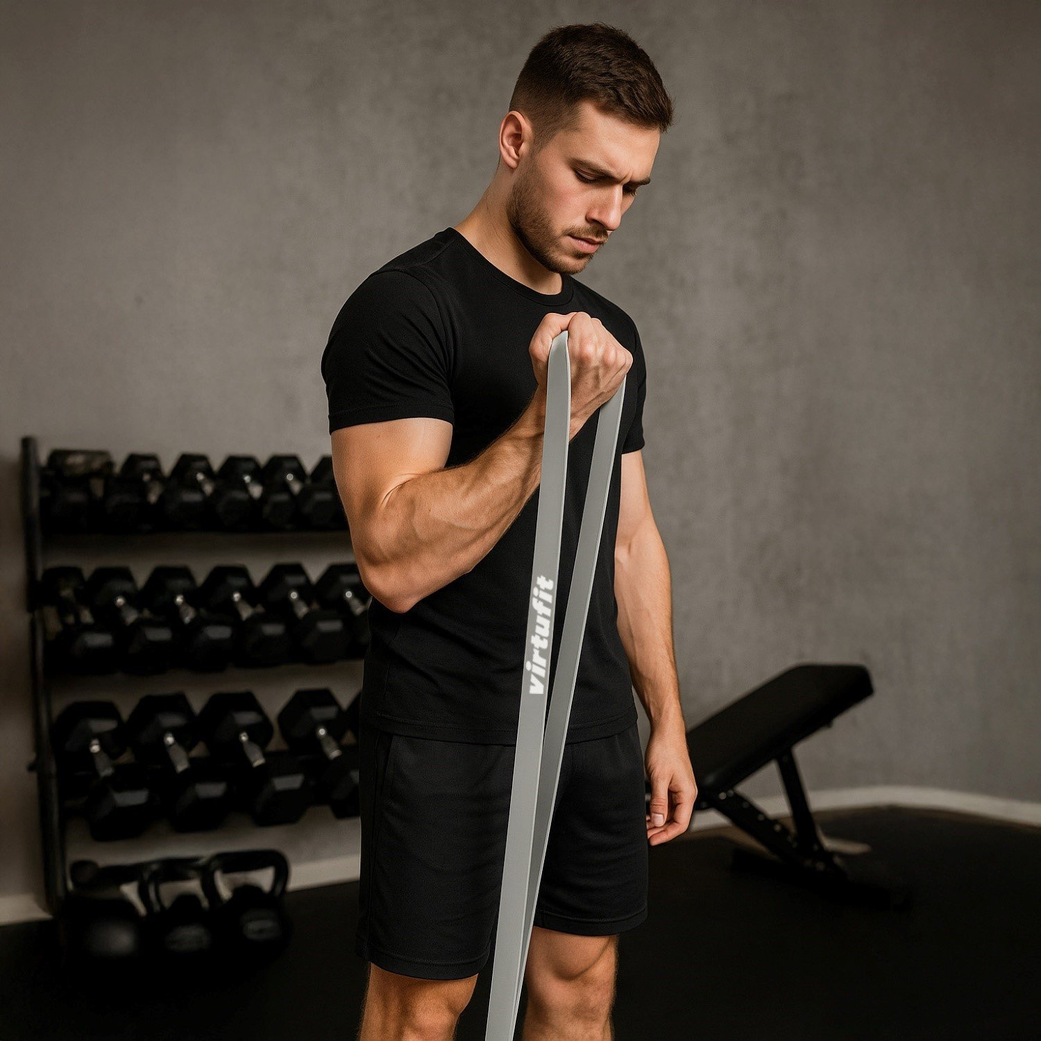 Ein Mann in schwarzer Kleidung trainiert mit einem grauen Widerstandsband. Hanteln und eine Bank sind im Hintergrund, Fitnessstudio.