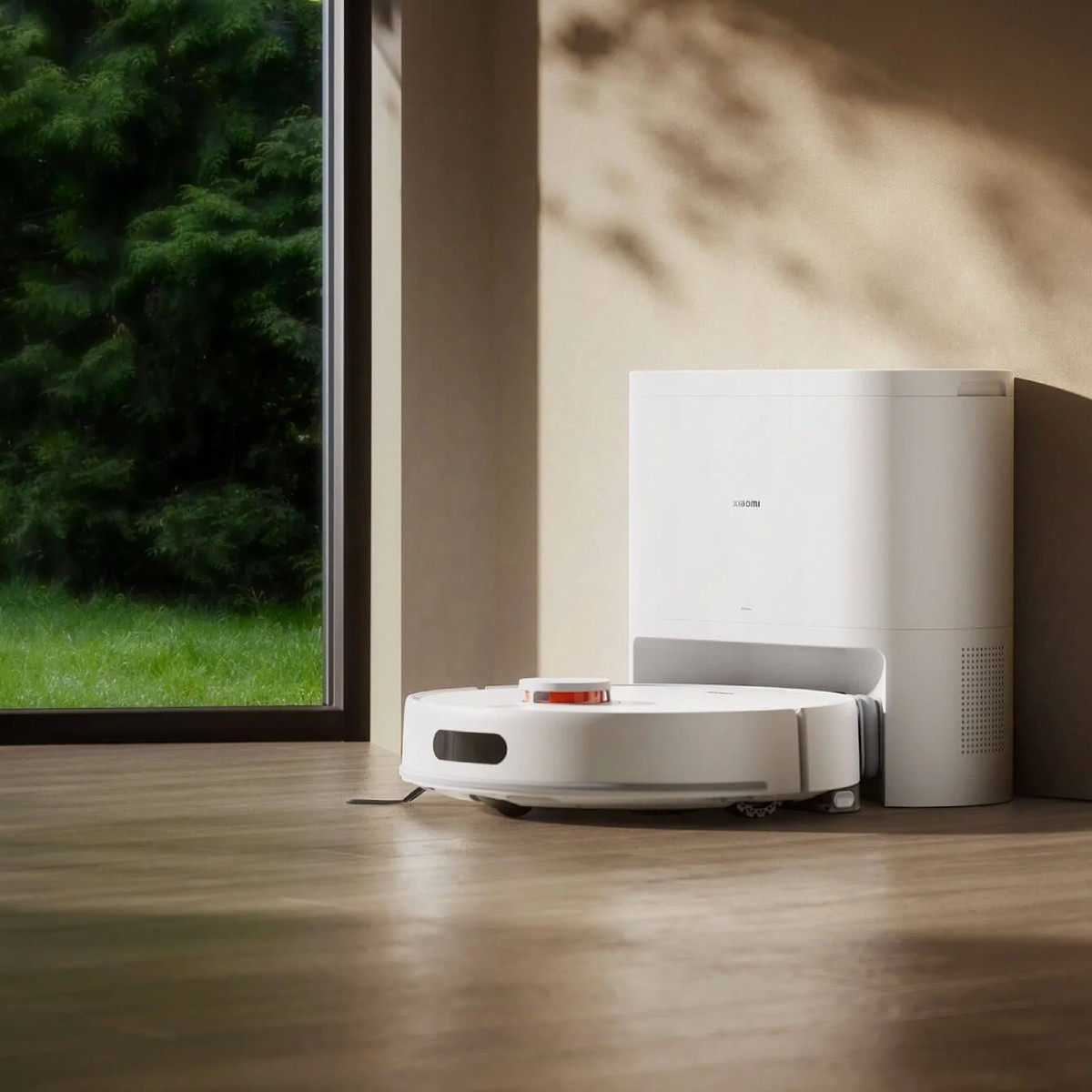 Een witte robotstofzuiger en een wit oplaadstation staan op een houten vloer. Een raam toont groene bomen buiten.