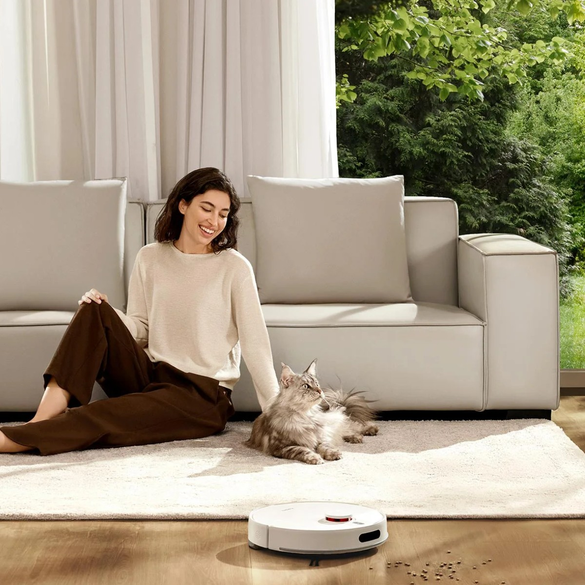 Vrouw en kat zitten bij een bank. Een robotstofzuiger staat op de voorgrond. Een raam met bomen op de achtergrond.