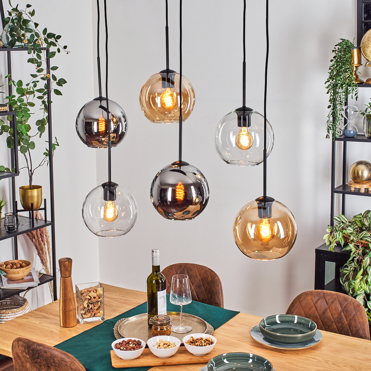 Esszimmer mit Holztisch, Hängelampen, Tellern, Schalen und Snacks. Pflanzen und Regale im Hintergrund.