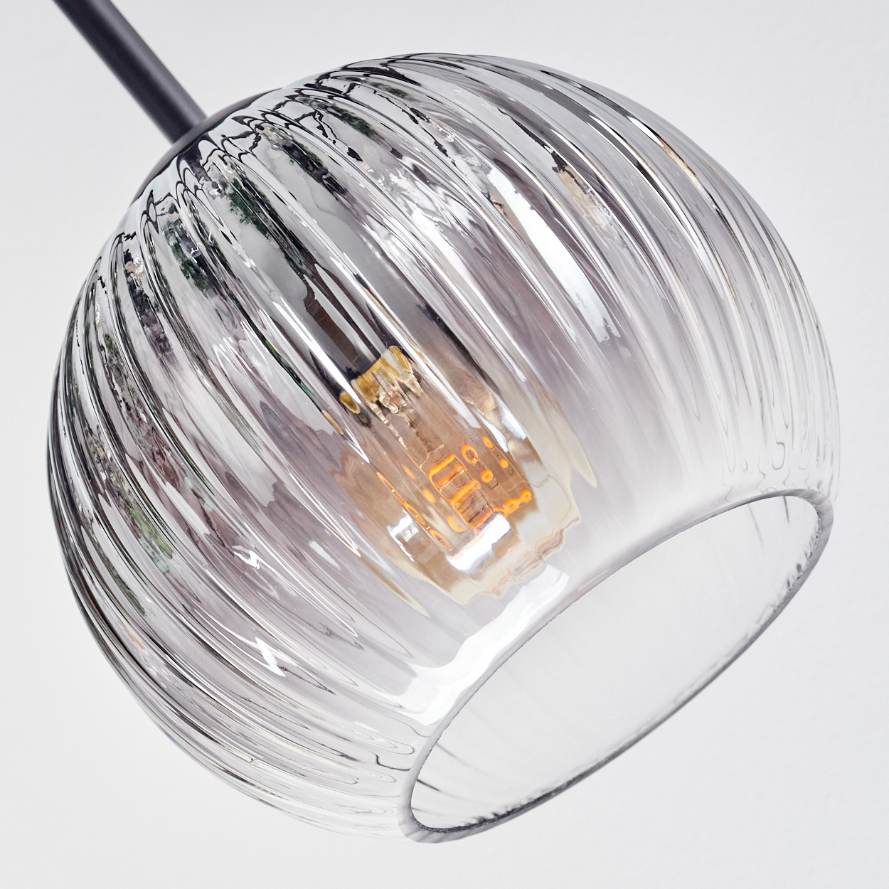 Eine klare, geriffelte Glaskugel-Hängelampe. Im Inneren leuchtet eine Glühbirne. Die Lampe hängt an einer schwarzen Metallstange vor weißem Hintergrund.