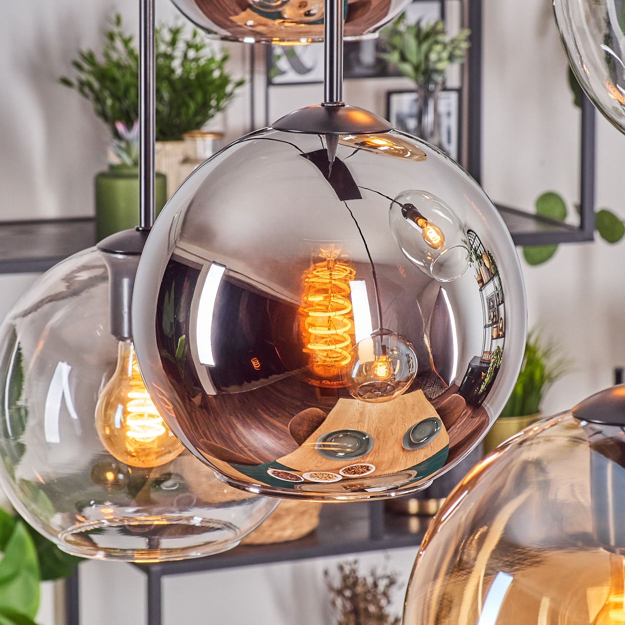 Nahaufnahme von modernen Pendelleuchten mit Glaskugeln. In den Kugeln spiegeln sich ein Tischgedeck und Pflanzen. Die Lichter haben warme, goldene Glühbirnen.