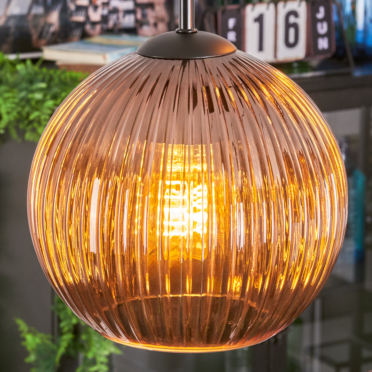 Ein runder, gerippter Glasschirm leuchtet mit warmem Licht. Der Hintergrund umfasst grüne Pflanzen und einen Kalender mit der Zahl 16.