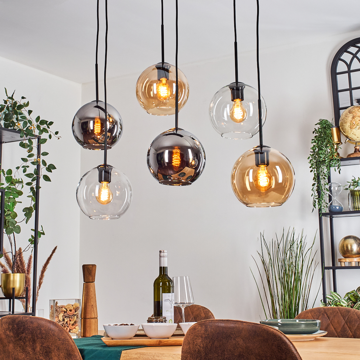 Esszimmer mit hängenden Kugelleuchten. Tisch mit Wein, Gläsern und Schalen. Pflanzen und Regale im Hintergrund.