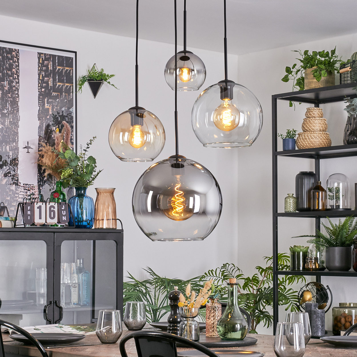 Esszimmer mit Hängelampen, Tisch, Pflanzen und schwarzen Metallregalen mit Dekoration.