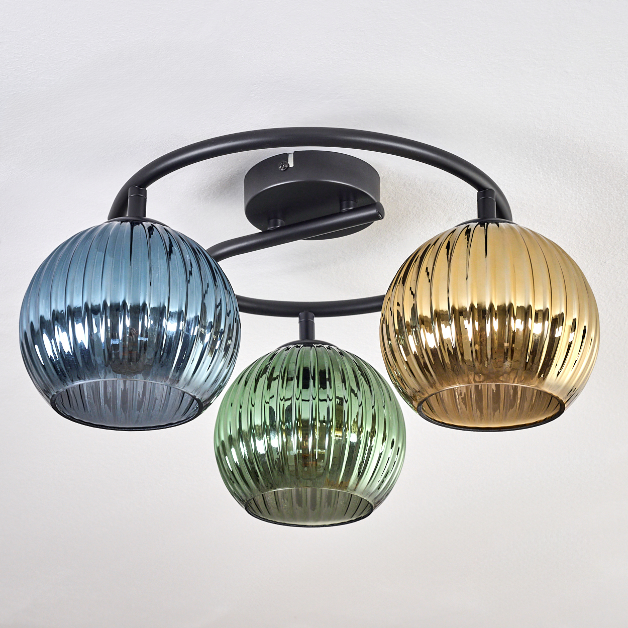 Eine Deckenleuchte mit drei Glasschirmen in Blau, Grün und Gold, montiert auf einem schwarzen Metallrahmen, vor weißer Decke.