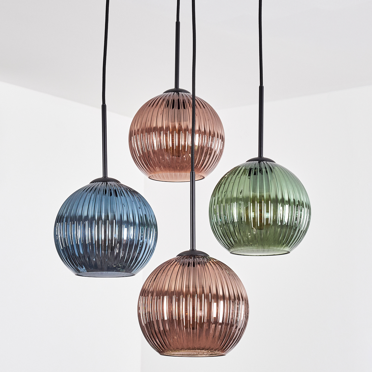 Vier bunte, gerippte Glas-Pendelleuchten hängen an schwarzen Kabeln von einer weißen Decke. Die Leuchten sind blau, grün und zwei Rosatöne.