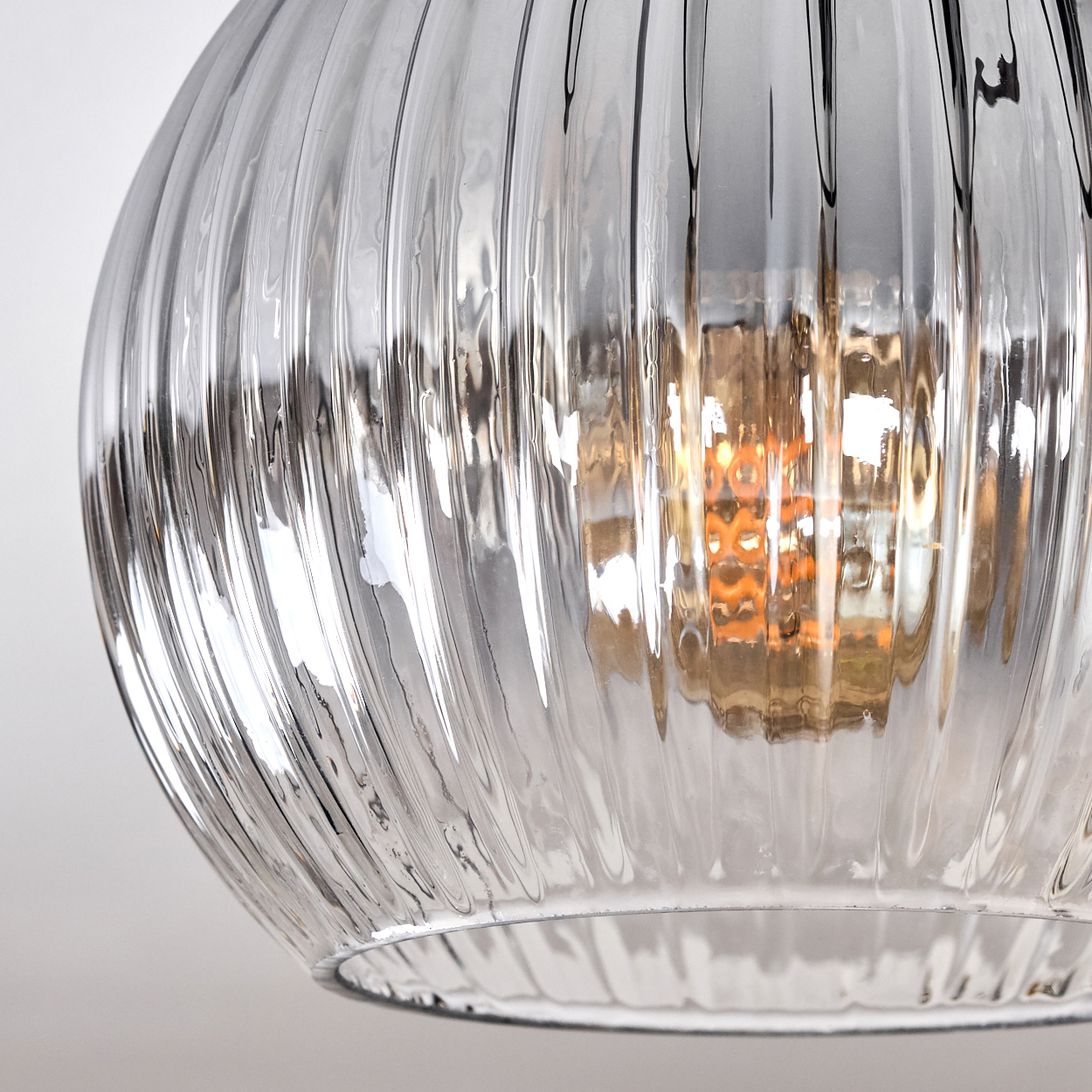 Nahaufnahme eines klaren, gerippten Glasschirmes. Im Inneren glüht eine warme Glühbirne. Der Hintergrund ist ein sanftes Weiß, das die Details der Lampe hervorhebt.