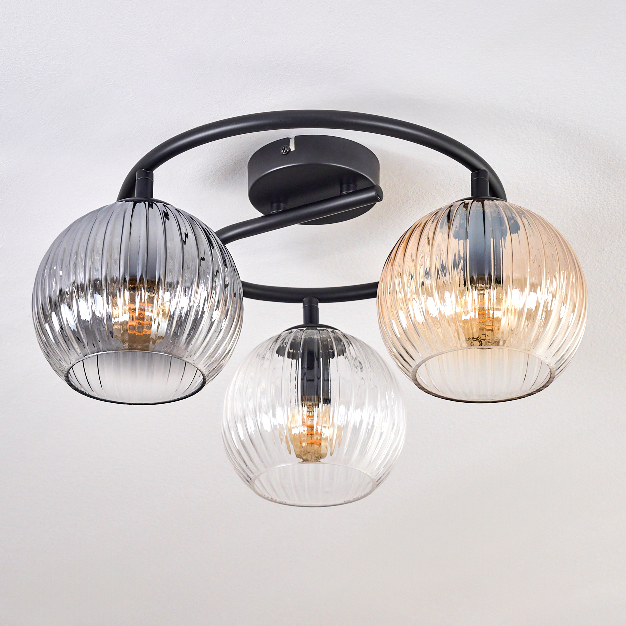 Moderne Deckenleuchte mit drei Glasschirmen. Die Schirme sind gerippt, in Silber, klar und Gold. Schwarzer Metallrahmen an einer weißen Decke.