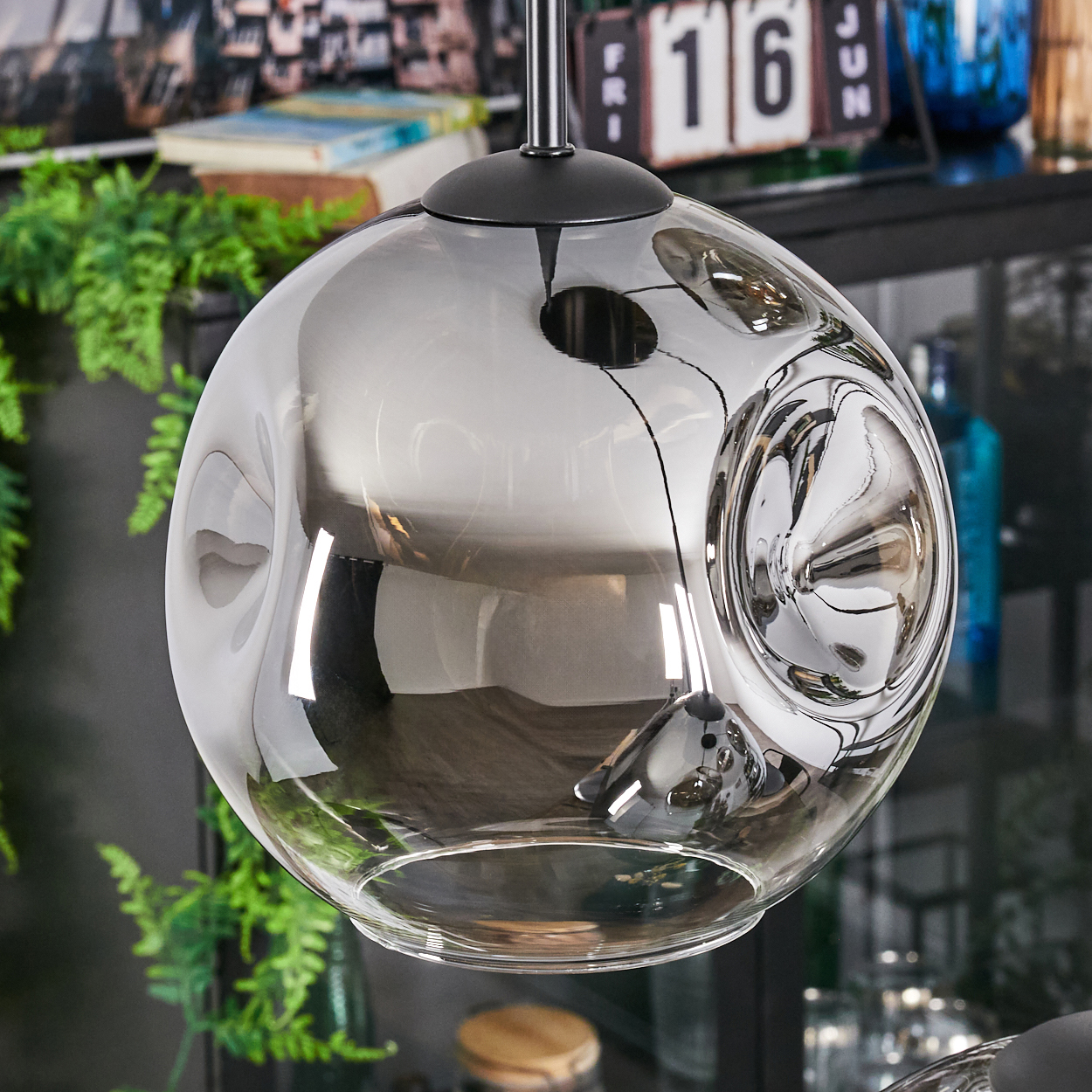 Eine moderne, kugelförmige Glaslampe hängt vor einer grünen Pflanze. Die Lampe spiegelt den Hintergrund wider, einschließlich eines Kalenders.