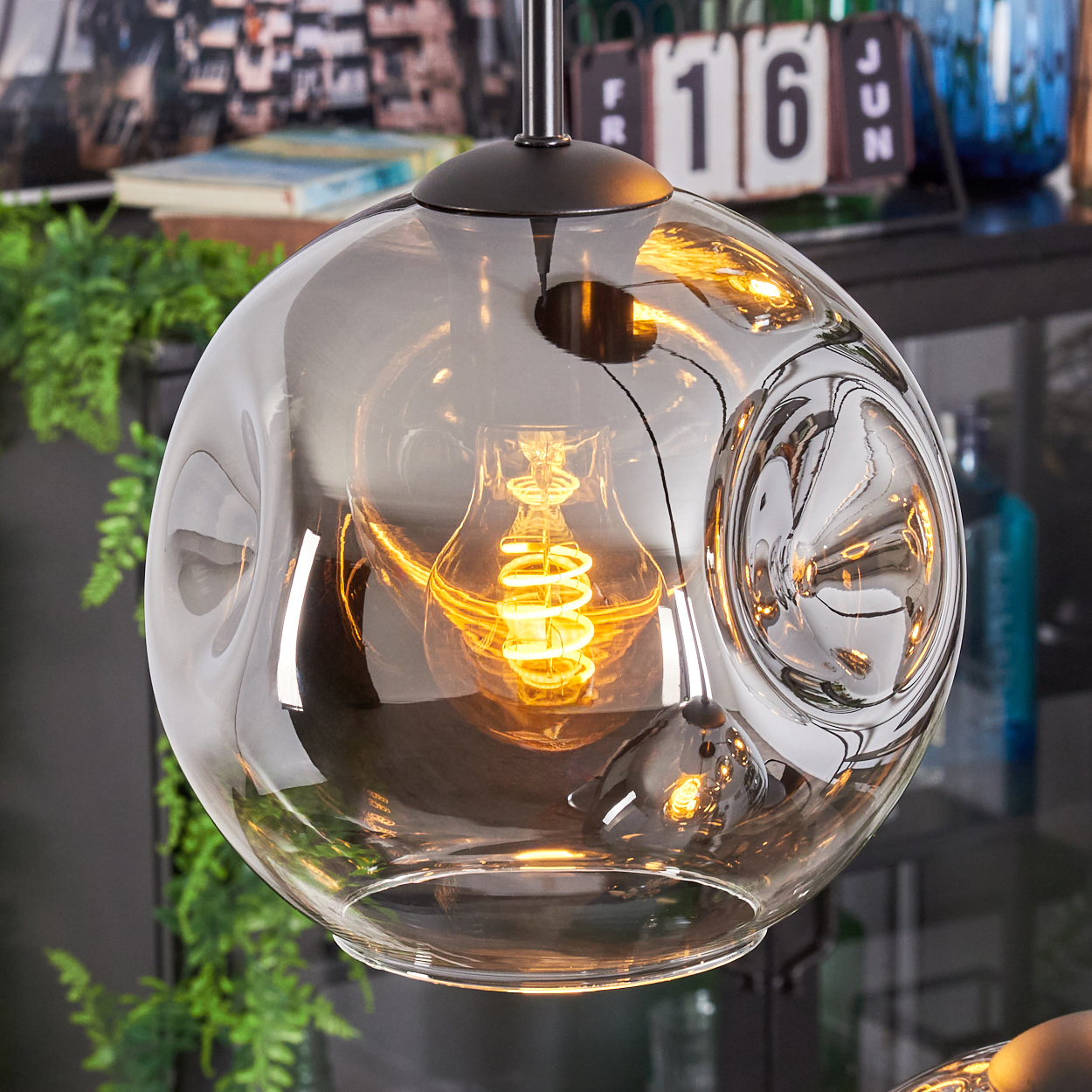 Eine moderne Glaskugellampe mit einer Wendelglühbirne. Die Lampe hängt von der Decke, mit einem Kalender und Pflanzen im Hintergrund.