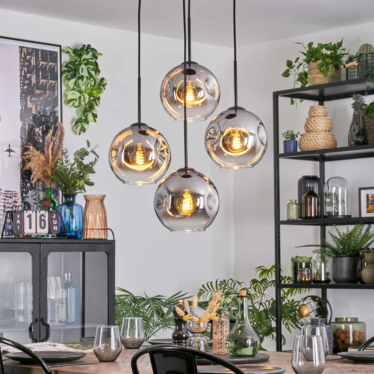 Vier kugelförmige Pendelleuchten hängen über einem Esstisch. Der Raum bietet Pflanzen, Dekorationen und ein schwarzes Metallregal.