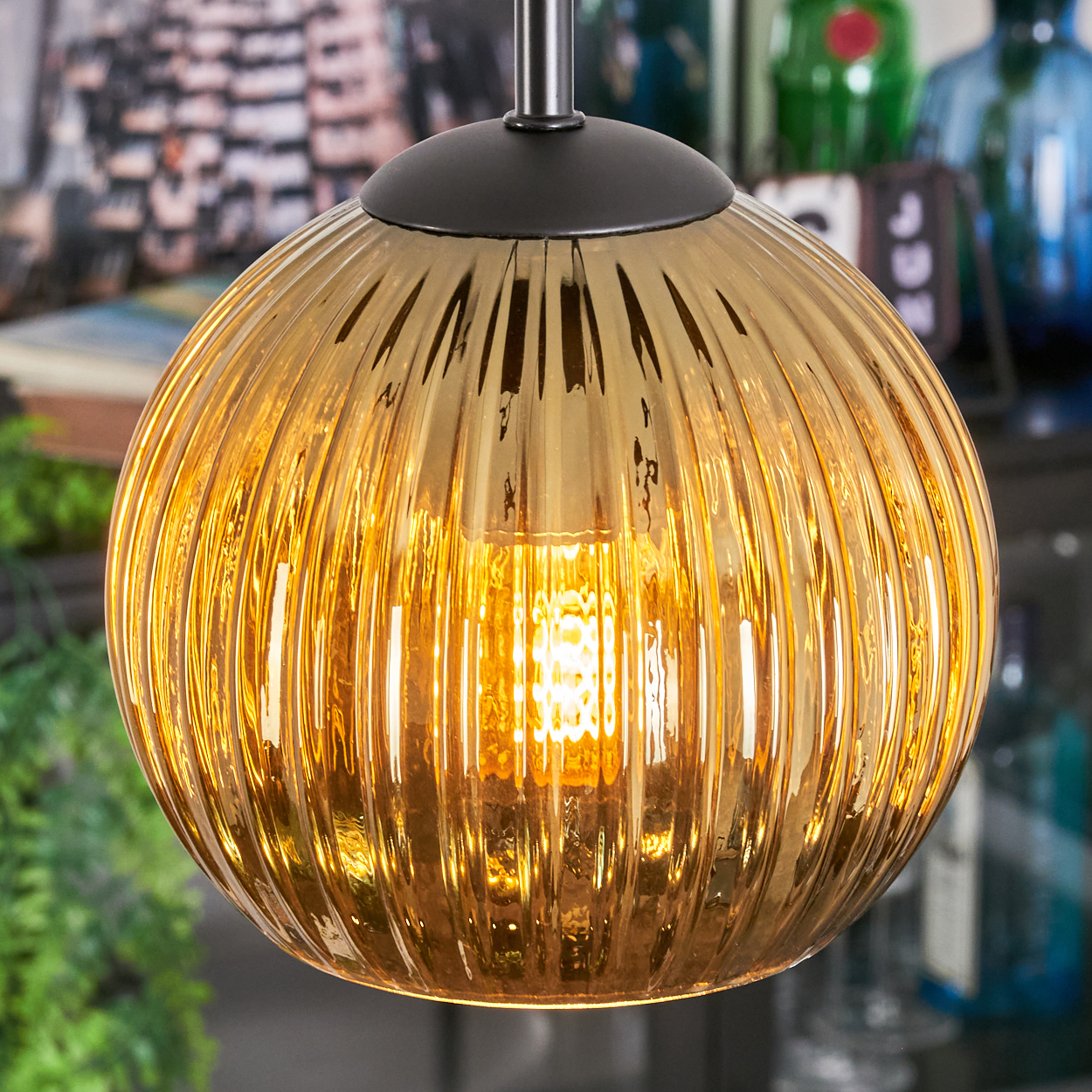 Eine goldene, gerippte Glas-Pendelleuchte hängt. Die Glühbirne im Inneren leuchtet hell. Der Hintergrund enthält eine verschwommene Szene mit Grün und Flaschen.