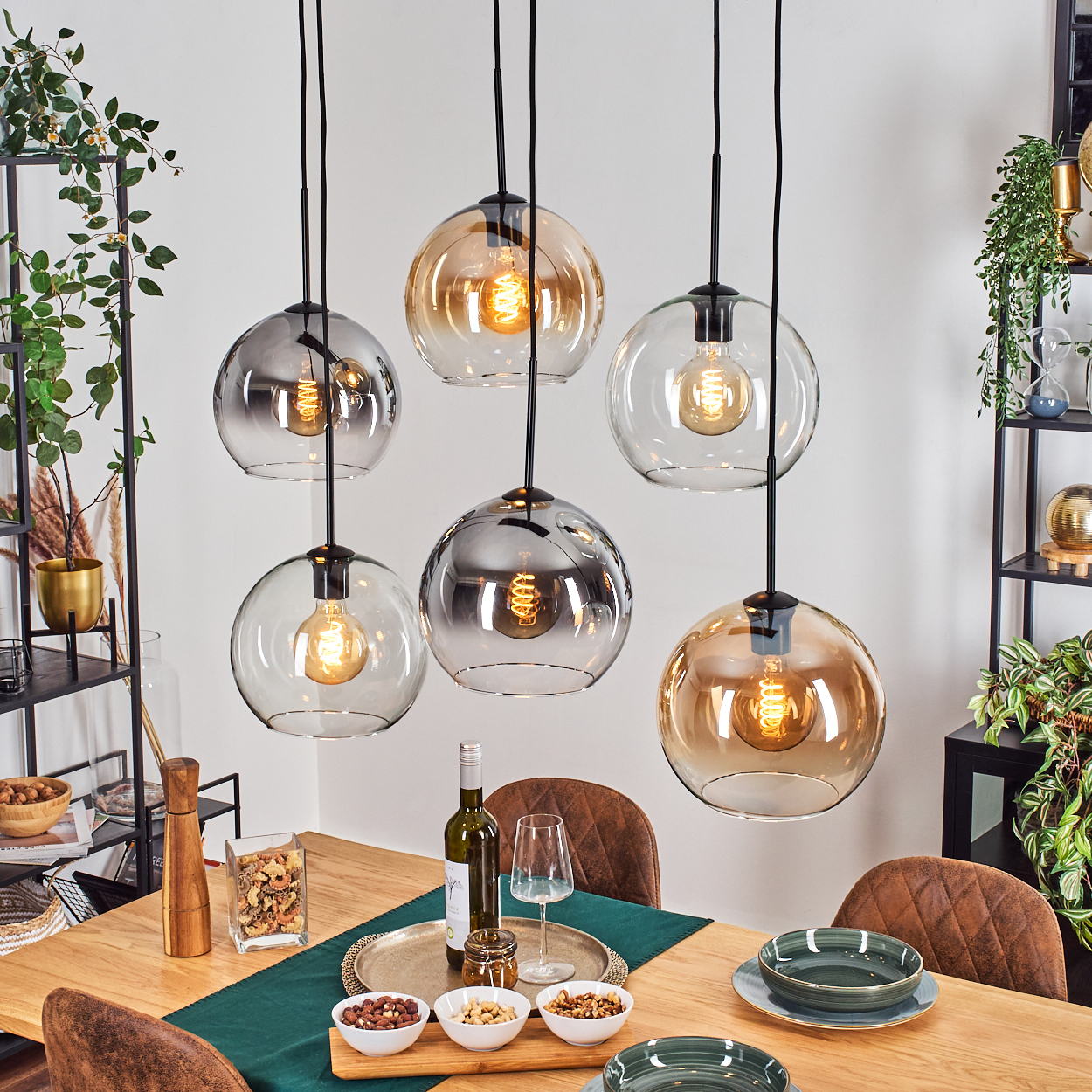 Esszimmer mit hängenden Kugelleuchten, Holztisch, Essen, Wein und Stühlen. Pflanzen und Regale im Hintergrund.