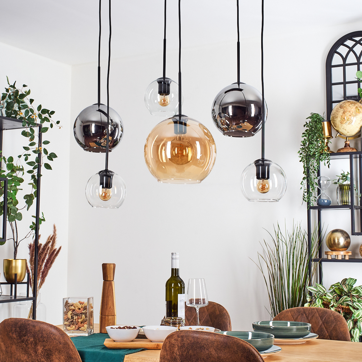Esszimmer mit Pendelleuchten, Tisch mit Wein, Gläsern und Schüsseln. Pflanzen und Regale im Hintergrund.