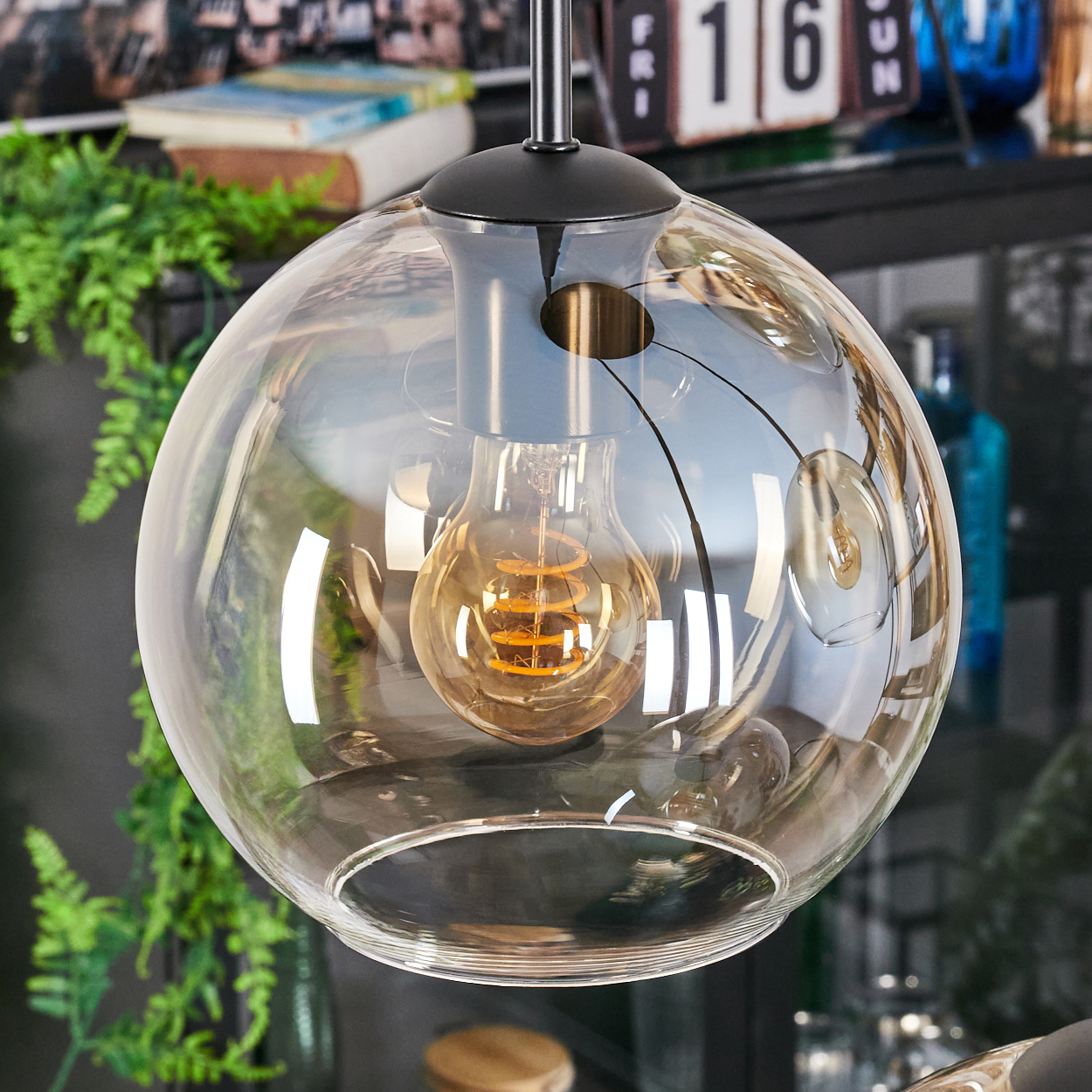 Eine klare Glaskugel-Pendelleuchte mit einer leuchtenden, warm getönten Glühbirne im Inneren. Im Hintergrund Pflanzen, ein Kalender und andere Dekorationen.